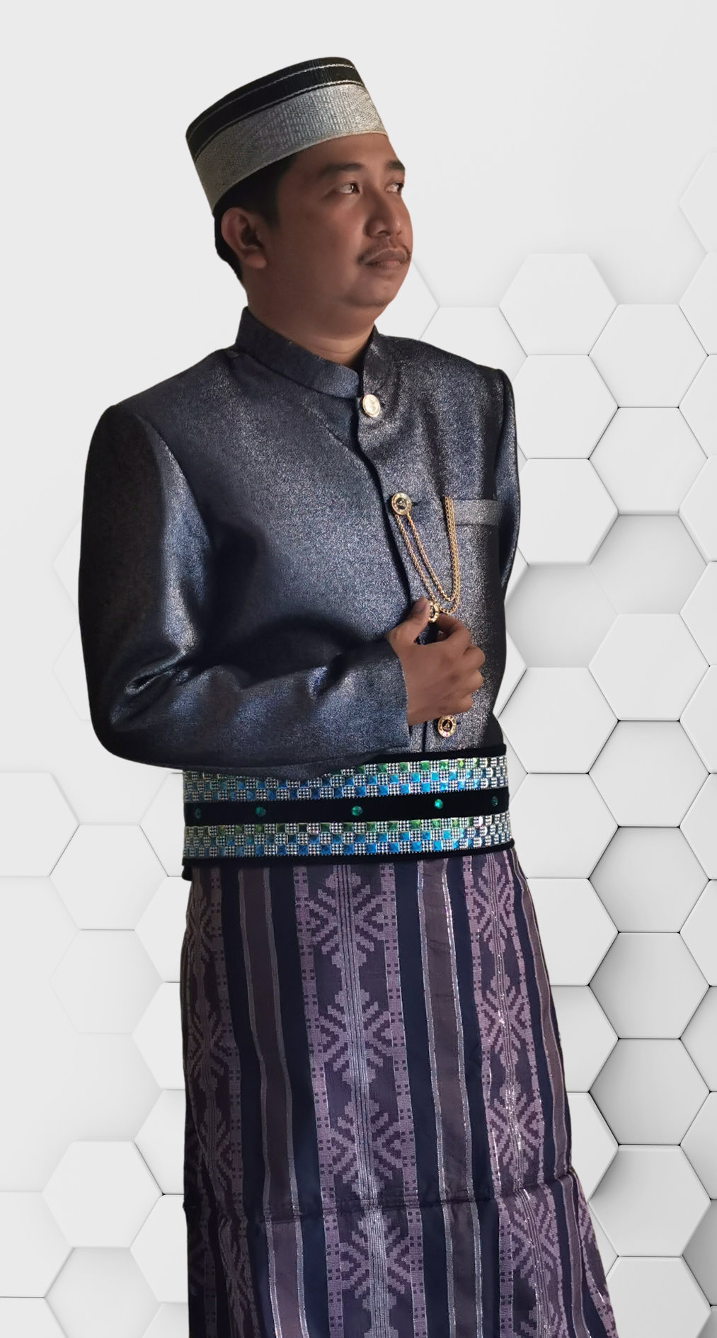 Photograph of a man wearing traditional formal attire. He is dressed in a dark metallic jacket with gold buttons and a decorative chain. The outfit includes a patterned sarong with shades of purple and blue. He is also wearing a traditional hat with a silver and black pattern. The background consists of a white hexagonal pattern.