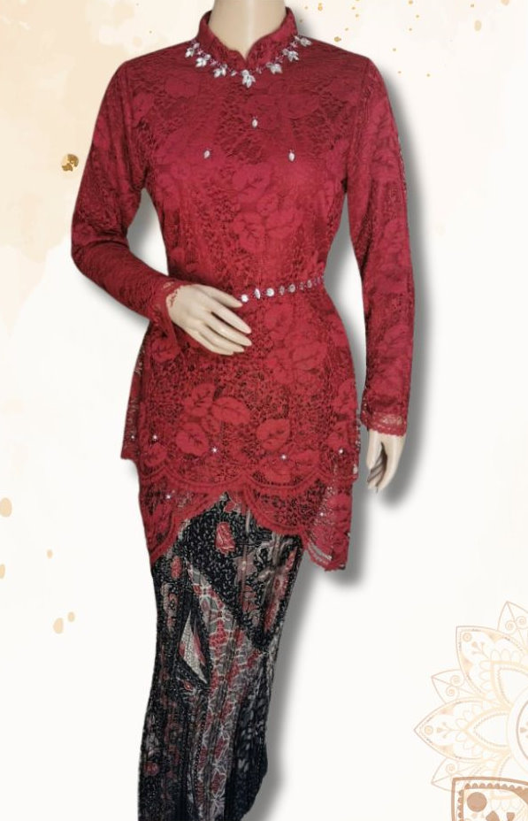 Photograph of a mannequin dressed in a traditional red lace kebaya. The kebaya features intricate floral patterns and is adorned with small decorative stones. It has a high neckline and long sleeves. The kebaya is paired with a black and red batik sarong featuring a detailed pattern. The background is light with subtle decorative elements